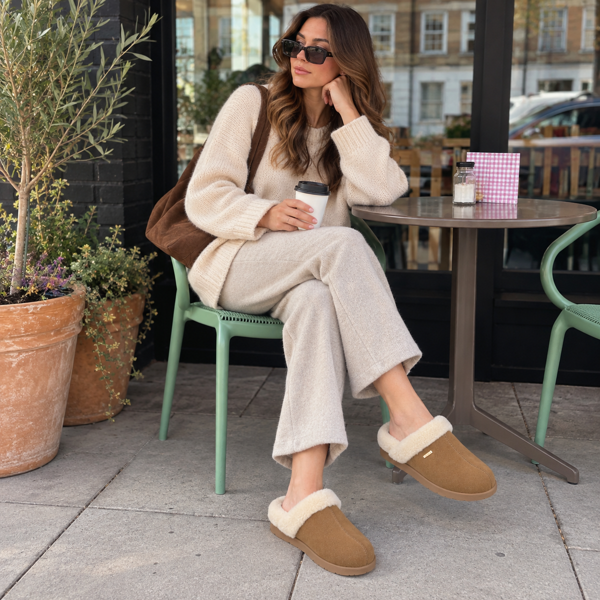 A woman sits at an outdoor cafe table wearing PCSsole brown slip-on slippers with white faux-fur trim, and a brown shoulder bag.