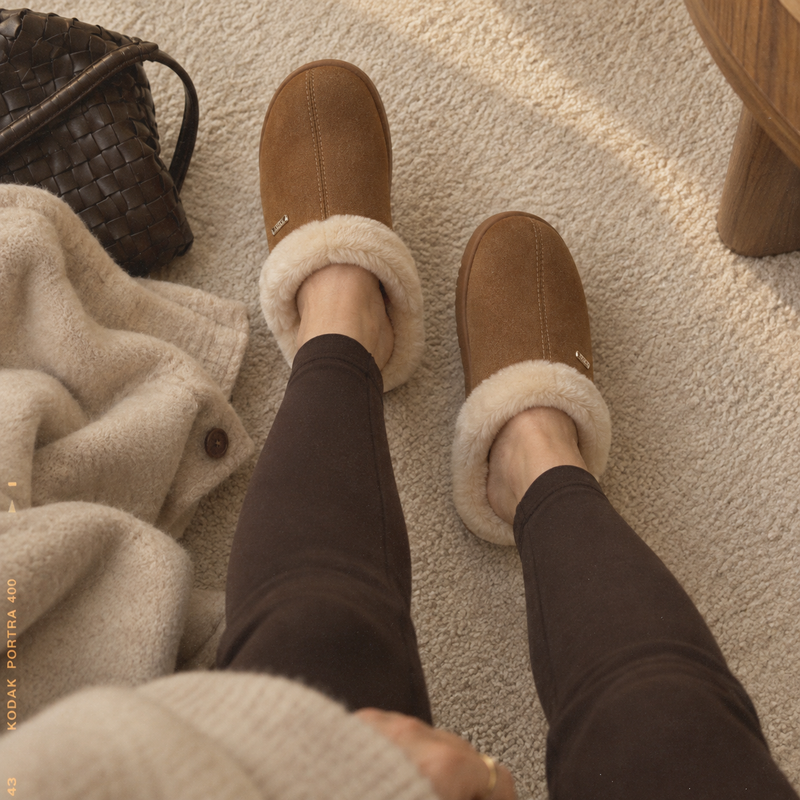 A woman wearing PCSsole brown suede slip-on slippers with fluffy cream lining