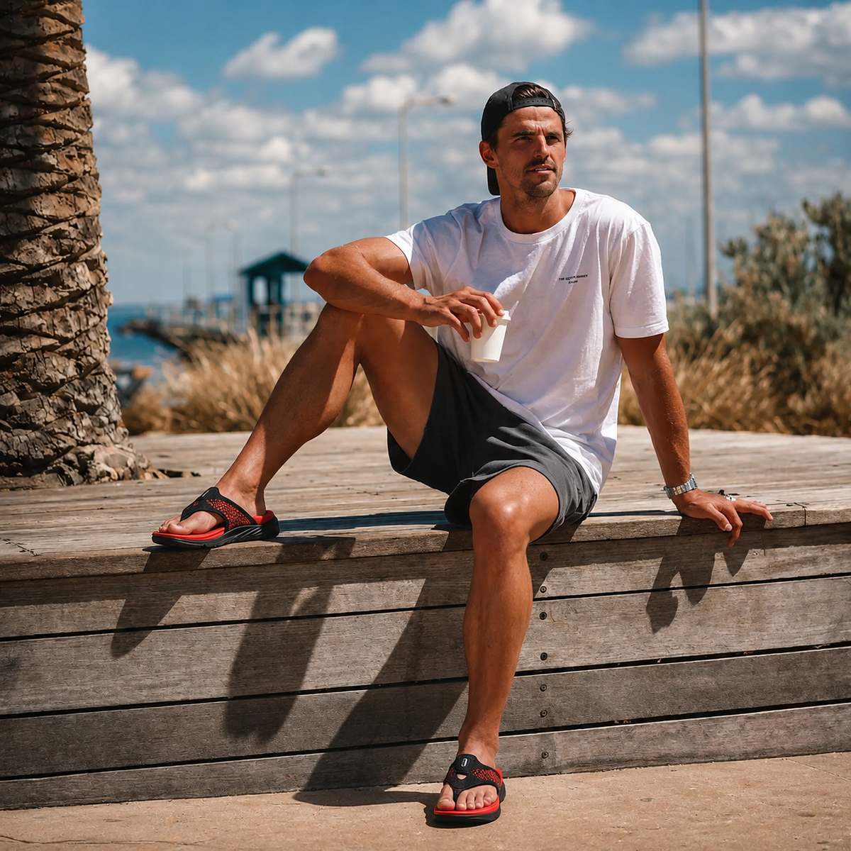 A man sitting on a wooden bench by a palm tree, wearing PCSsole crimson red flip flops.