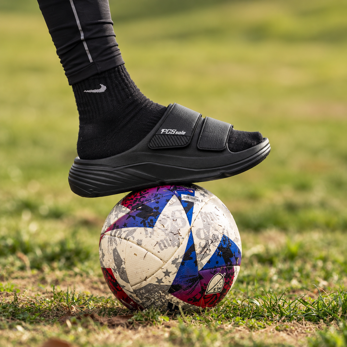 A man in thick-soled PCSsole adjustable recovery slides, resting his foot on a colorful soccer ball on the grass.